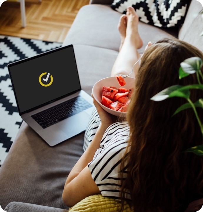 一個女人躺在沙發上吃一碗水果，旁邊有一台畫面上顯示 Norton 標誌的筆記型電腦